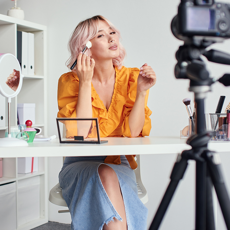 Young beautiful blond asian woman is recording make up tutorial video for beauty blog, wearing terracota blouse. Video blogging, isolation, stay home concept - Instagram Reels for Brands