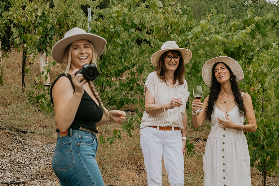 Behind the Scenes of our photoshoot with Vindemia Winery. Photographer holding Canon Mirrorless Camera in the vineyards smiling with two models in their 60's holding wine for a winery photoshoot.